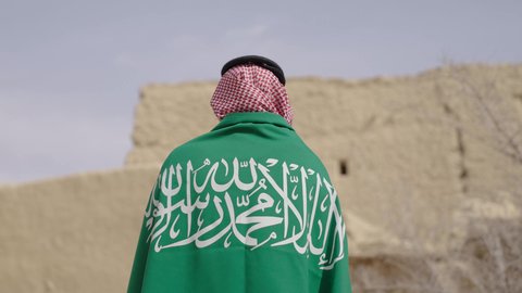 Love of the homeland and belonging to it, the anniversary of the founding of the first Saudi state 1727 AD, a picture from the back of an elderly Saudi Arabian Gulf man wearing a traditional shemagh and thobe holding the national flag in his hand to celebrate Flag Day, March 11, a walk in an old traditional popular place, the day of our beginning, February 22