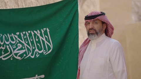 مفهوم حب الوطن والانتماء اليه, ذكرى تأسيس الدولة السعودية الأولى1727 م, رجل كبير بالسن عربي خليجي سعودي يرتدي الشماغ والثوب التقليدي يمسك بيده راية الوطن للاحتفال بيوم العلم 11 مارس، التجول في مكان شعبي تقليدي قديم، يوم بدينا 22 فبراير