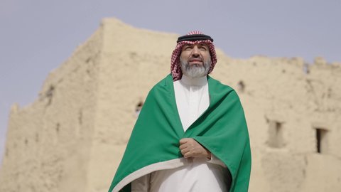 The concept of patriotism and belonging to the homeland, the anniversary of the founding of the first Saudi state 1727 AD, an elderly Saudi Gulf Arab man wearing a traditional shemagh and thobe holding the national flag in his hand to celebrate Flag Day, March 11, a walk in an old traditional popular place, the day of our beginning, February 22
