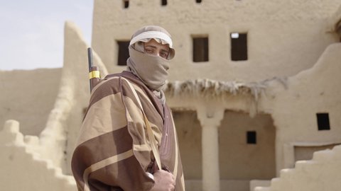 Preserving and defending the history and heritage of the Kingdom, the day we began, February 22, war and fighting with guns to protect the homeland, the establishment of the first Saudi state in 1727 AD, a Saudi Gulf Arab man wearing traditional folk costume carrying a gun in a mud-brick neighborhood, the concept of surveillance and guarding