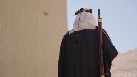 Defending and preserving the history and heritage of the Kingdom, the day we began, February 22, war and fighting with guns to protect the homeland, the establishment of the first Saudi state in 1727 AD, a Saudi Gulf Arab man wearing traditional folk costume carrying a gun in a mud-brick neighborhood, the concept of surveillance and guarding