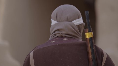 الحرب والقتال بالبنادق لحماية الوطن، تأسيس الدولة السعودية الأولى 1727 م، رجل عربي خليجي سعودي يرتدي الزي الشعبي التقليدي يحمل البندقية في احياء شعبية طينية، مفهوم المراقبة والحراسة، الحفاظ والدفاع على تاريخ وتراث المملكة، يوم بدينا 22 فبراير