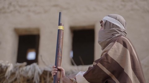 الحرب والقتال بالبنادق لحماية الوطن، تأسيس الدولة السعودية الأولى 1727 م، رجل عربي خليجي سعودي يرتدي الزي الشعبي التقليدي يحمل البندقية في احياء شعبية طينية، مفهوم المراقبة والحراسة، الحفاظ والدفاع على تاريخ وتراث المملكة، يوم بدينا 22 فبراير