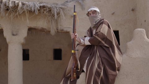 الحرب والقتال بالبنادق لحماية الوطن، تأسيس الدولة السعودية الأولى 1727 م، رجل عربي خليجي سعودي يرتدي الزي الشعبي التقليدي يحمل البندقية في احياء شعبية طينية، مفهوم المراقبة والحراسة، الحفاظ والدفاع على تاريخ وتراث المملكة، يوم بدينا 22 فبراير