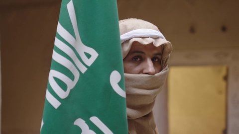 ارتداء الزي الشعبي بالمناسبة الوطنية، يوم بدينا 22 فبراير, حب الوطن والانتماء اليه, رجل عربي خليجي سعودي يرتدي البشت والغترة يقف في منزل طيني بجانب علم يوم التأسيس بتعابير الفخر والاعتزاز، الاحتفال بذكرى تأسيس الدولة السعودية الأولى 1727 م