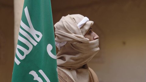 الاحتفال بذكرى تأسيس الدولة السعودية الأولى 1727 م, ارتداء الزي الشعبي بالمناسبة الوطنية، يوم بدينا 22 فبراير, حب الوطن والانتماء اليه, رجل عربي خليجي سعودي يرتدي البشت والغترة يقف في منزل طيني بجانب علم يوم التأسيس بتعابير الفخر والاعتزاز