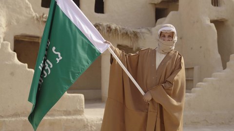 ارتداء الزي الشعبي بالمناسبة الوطنية، يوم بدينا 22 فبراير, حب الوطن والانتماء اليه, رجل عربي خليجي سعودي يرتدي البشت والغترة يقف في منزل طيني بجانب علم يوم التأسيس بتعابير الفخر والاعتزاز، الاحتفال بذكرى تأسيس الدولة السعودية الأولى 1727 م