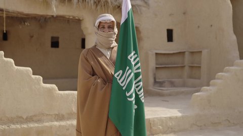Celebrating the anniversary of the founding of the first Saudi state in 1727 AD, wearing traditional costume on the national occasion, the day of our beginning, February 22, love of the homeland and belonging to it, a Saudi Gulf Arab man wearing a bisht and ghutra stands in a mud house next to the flag of the founding day with expressions of pride and honor