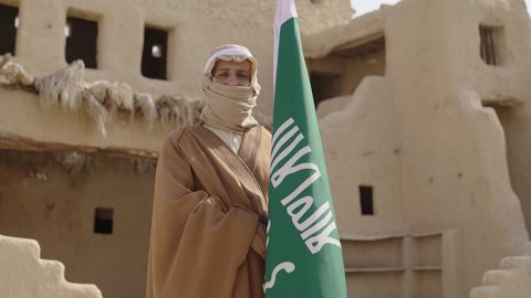 الاحتفال بذكرى تأسيس الدولة السعودية الأولى 1727 م, ارتداء الزي الشعبي بالمناسبة الوطنية، يوم بدينا 22 فبراير, حب الوطن والانتماء اليه, رجل عربي خليجي سعودي يرتدي البشت والغترة يقف في منزل طيني بجانب علم يوم التأسيس بتعابير الفخر والاعتزاز