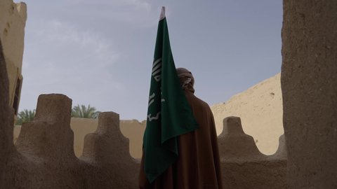 الاحتفال بذكرى تأسيس الدولة السعودية الأولى 1727 م, ارتداء الزي الشعبي بالمناسبة الوطنية، يوم بدينا 22 فبراير, حب الوطن والانتماء اليه, رجل عربي خليجي سعودي يرتدي البشت والغترة يقف في منزل طيني بجانب علم يوم التأسيس بتعابير الفخر والاعتزاز