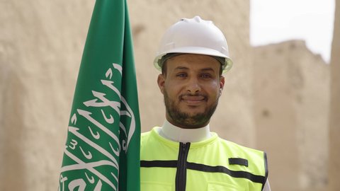 Raising the Kingdom's flag in popular heritage sites on March 11, ancient architecture and mud house construction, a smiling Saudi Gulf Arab engineer wearing a jacket and protective helmet with gestures of pride and honor on National Day, September 23, the day we began in 1727 AD, the anniversary of the founding of the first Saudi state, February 22
