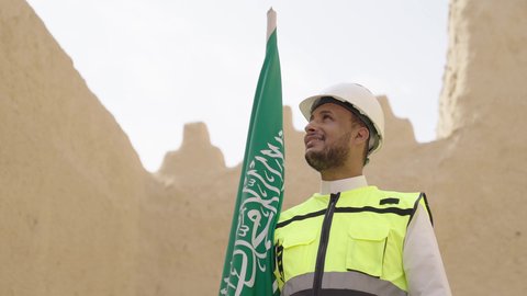 رفع علم المملكة في الأماكن التراثيه الشعبيه في 11 مارس، فن العمارة القديم وبناء البيوت الطينية، مهندس عربي خليجي سعودي مبتسم يرتدي السترة وخوذة الحماية بايماءات الفخر والاعتزاز في اليوم الوطني 23 سبتمبر، يوم بدينا 1727 م، ذكرى تأسيس الدولة السعودية الأولى 22 فبراير