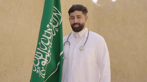 يوم بدينا 1727 م، مهن ووظائف سعودية طبية، مفهوم العمل في القطاع الطبي، طبيب عربي خليجي سعودي يرتدي الثوب التقليدي والزي الطبي والسماعات يقف في بيت طيني ويحمل بيده علم المملكة، إحياء ذكرى اليوم الوطني السعودي 23سبتمبر، الإحتفال بيوم العلم 11 مارس