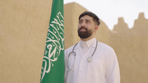 يوم بدينا 1727 م، مهن ووظائف سعودية طبية، مفهوم العمل في القطاع الطبي، طبيب عربي خليجي سعودي يرتدي الثوب التقليدي والزي الطبي والسماعات يقف في بيت طيني ويحمل بيده علم المملكة، إحياء ذكرى اليوم الوطني السعودي 23سبتمبر، الإحتفال بيوم العلم 11 مارس