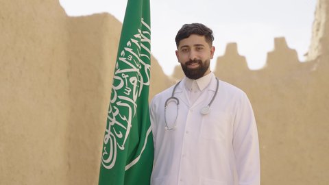 يوم بدينا 1727 م، مهن ووظائف سعودية طبية، مفهوم العمل في القطاع الطبي، طبيب عربي خليجي سعودي يرتدي الثوب التقليدي والزي الطبي والسماعات يقف في بيت طيني ويحمل بيده علم المملكة، إحياء ذكرى اليوم الوطني السعودي 23سبتمبر، الإحتفال بيوم العلم 11 مارس