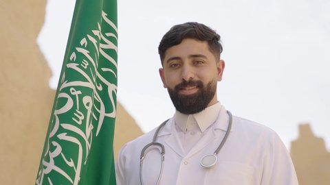 وظائف ومهن سعودية طبية، مفهوم العمل في القطاع الطبي، طبيب عربي خليجي سعودي يرتدي الثوب التقليدي والزي الطبي والسماعات يقف في بيت طيني ويحمل بيده علم المملكة، إحياء ذكرى اليوم الوطني السعودي 23سبتمبر، الإحتفال بيوم العلم 11 مارس