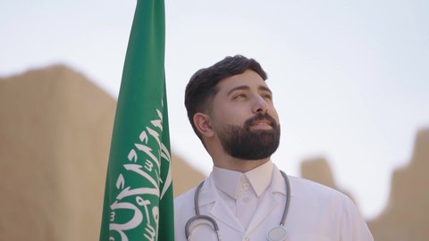 مهن ووظائف سعودية طبية، مفهوم العمل في القطاع الطبي، طبيب عربي خليجي سعودي يرتدي الثوب التقليدي والزي الطبي والسماعات يقف في بيت طيني ويحمل بيده علم المملكة، إحياء ذكرى اليوم الوطني السعودي 23سبتمبر، الإحتفال بيوم العلم 11 مارس