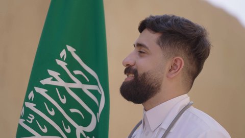 مهن ووظائف سعودية طبية، مفهوم العمل في القطاع الطبي، طبيب عربي خليجي سعودي يرتدي الثوب التقليدي والزي الطبي والسماعات يقف في بيت طيني ويحمل بيده علم المملكة، إحياء ذكرى اليوم الوطني السعودي 23سبتمبر، الإحتفال بيوم العلم 11 مارس