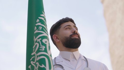 مهن ووظائف سعودية طبية، مفهوم العمل في القطاع الطبي، طبيب عربي خليجي سعودي يرتدي الثوب التقليدي والزي الطبي والسماعات يقف في بيت طيني ويحمل بيده علم المملكة، إحياء ذكرى اليوم الوطني السعودي 23سبتمبر، الإحتفال بيوم العلم 11 مارس