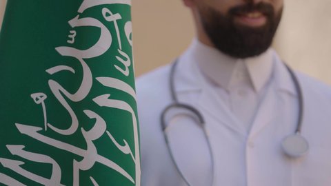 مهن ووظائف سعودية طبية، مفهوم العمل في القطاع الطبي، طبيب عربي خليجي سعودي يرتدي الثوب التقليدي والزي الطبي والسماعات يقف في بيت طيني ويحمل بيده علم المملكة، إحياء ذكرى اليوم الوطني السعودي 23سبتمبر، الإحتفال بيوم العلم 11 مارس
