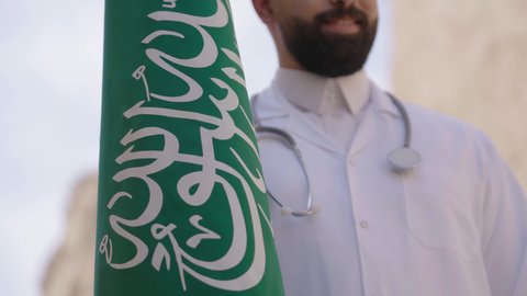 مهن ووظائف سعودية طبية، مفهوم العمل في القطاع الطبي، طبيب عربي خليجي سعودي يرتدي الثوب التقليدي والزي الطبي والسماعات يقف في بيت طيني ويحمل بيده علم المملكة، إحياء ذكرى اليوم الوطني السعودي 23سبتمبر، الإحتفال بيوم العلم 11 مارس