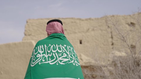 بناء البيوت الطينية وفن العمارة القديم، يوم العلم 11 مارس, إحياء ذكرى اليوم الوطني السعودي 23 سبتمبر, إيماءات الفخر و السرور بالانتماء للوطن, رجل عربي خليجي سعودي مبتسم يرتدي الشماغ و الثوب التقليدي يقف في بيت طيني ويحمل بيده علم المملكة، يوم بدينا 1727 م