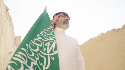 بناء البيوت الطينية وفن العمارة القديم، يوم العلم 11 مارس, إحياء ذكرى اليوم الوطني السعودي 23 سبتمبر, إيماءات الفخر و السرور بالانتماء للوطن, رجل عربي خليجي سعودي مبتسم يرتدي الشماغ و الثوب التقليدي يقف في بيت طيني ويحمل بيده علم المملكة، يوم بدينا 1727 م