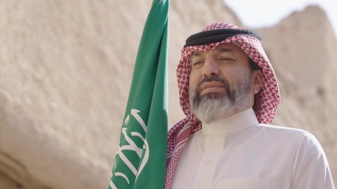 بناء البيوت الطينية وفن العمارة القديم، يوم العلم 11 مارس, إحياء ذكرى اليوم الوطني السعودي 23 سبتمبر, إيماءات الفخر و السرور بالانتماء للوطن, رجل عربي خليجي سعودي مبتسم يرتدي الشماغ و الثوب التقليدي يقف في بيت طيني ويحمل بيده علم المملكة، يوم بدينا 1727 م