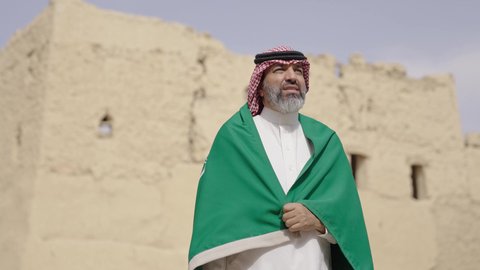 Ancient architecture and mud house construction, Flag Day March 11, Saudi National Day commemoration September 23, gestures of pride and joy in belonging to the homeland, a smiling Saudi Arabian Gulf man wearing a traditional shemagh and thobe standing in a mud house holding the Kingdm's flag, the day we began 1727 AD