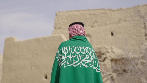 فن العمارة القديم وبناء البيوت الطينية، يوم العلم 11 مارس, إحياء ذكرى اليوم الوطني السعودي 23 سبتمبر, إيماءات الفخر و السرور بالانتماء للوطن, رجل عربي خليجي سعودي مبتسم يرتدي الشماغ و الثوب التقليدي يقف في بيت طيني ويحمل بيده علم المملكة، يوم بدينا 1727 م