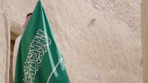 إيماءات الفخر و السرور بالانتماء للوطن, رجل عربي خليجي سعودي مبتسم يرتدي الشماغ و الثوب التقليدي يقف في بيت طيني ويحمل بيده علم المملكة، يوم بدينا 1727 م، فن العمارة القديم وبناء البيوت الطينية، يوم العلم 11 مارس, إحياء ذكرى اليوم الوطني السعودي 23 سبتمبر