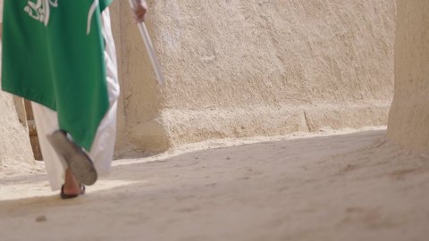 إيماءات الفخر و السرور بالانتماء للوطن, رجل عربي خليجي سعودي مبتسم يرتدي الشماغ و الثوب التقليدي يقف في بيت طيني ويحمل بيده علم المملكة، يوم بدينا 1727 م، فن العمارة القديم وبناء البيوت الطينية، يوم العلم 11 مارس, إحياء ذكرى اليوم الوطني السعودي 23 سبتمبر