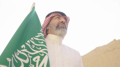 إيماءات الفخر و السرور بالانتماء للوطن, رجل عربي خليجي سعودي مبتسم يرتدي الشماغ و الثوب التقليدي يقف في بيت طيني ويحمل بيده علم المملكة، يوم بدينا 1727 م، فن العمارة القديم وبناء البيوت الطينية، يوم العلم 11 مارس, إحياء ذكرى اليوم الوطني السعودي 23 سبتمبر