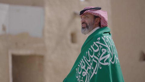 Ancient architecture and mud house construction, Flag Day March 11, Saudi National Day commemoration September 23, gestures of pride and joy in belonging to the homeland, a smiling Saudi Arabian Gulf man wearing a traditional shemagh and thobe standing in a mud house holding the Kingdom's flag, the day we began 1727 AD