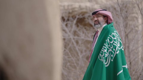 Ancient architecture and mud house construction, Flag Day March 11, Saudi National Day commemoration September 23, gestures of pride and joy in belonging to the homeland, a smiling Saudi Arabian Gulf man wearing a traditional shemagh and thobe standing in a mud house holding the Kingdom's flag, the day we began 1727 AD