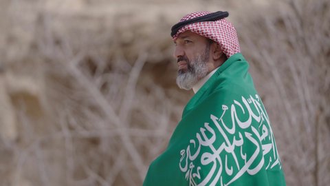 Ancient architecture and mud house construction, Flag Day March 11, Saudi National Day commemoration September 23, gestures of pride and joy in belonging to the homeland, a smiling Saudi Arabian Gulf man wearing a traditional shemagh and thobe standing in a mud house holding the Kingdom's flag, the day we began 1727 AD