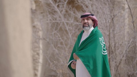 فن العمارة القديم وبناء البيوت الطينية، يوم العلم 11 مارس, إحياء ذكرى اليوم الوطني السعودي 23 سبتمبر, إيماءات الفخر و السرور بالانتماء للوطن, رجل عربي خليجي سعودي مبتسم يرتدي الشماغ و الثوب التقليدي يقف في بيت طيني ويحمل بيده علم المملكة، يوم بدينا 1727 م