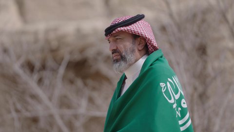 فن العمارة القديم وبناء البيوت الطينية، يوم العلم 11 مارس, إحياء ذكرى اليوم الوطني السعودي 23 سبتمبر, إيماءات الفخر و السرور بالانتماء للوطن, رجل عربي خليجي سعودي مبتسم يرتدي الشماغ و الثوب التقليدي يقف في بيت طيني ويحمل بيده علم المملكة، يوم بدينا 1727 م