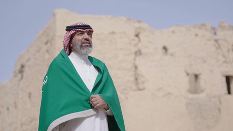 فن العمارة القديم وبناء البيوت الطينية، يوم العلم 11 مارس, إحياء ذكرى اليوم الوطني السعودي 23 سبتمبر, إيماءات الفخر و السرور بالانتماء للوطن, رجل عربي خليجي سعودي مبتسم يرتدي الشماغ و الثوب التقليدي يقف في بيت طيني ويحمل بيده علم المملكة، يوم بدينا 1727 م
