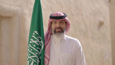 Ancient architecture and mud house construction, Flag Day March 11, Saudi National Day commemoration September 23, gestures of pride and joy in belonging to the homeland, a smiling Saudi Arabian Gulf man wearing a traditional shemagh and thobe standing in a mud house holding the Kingdom's flag, the day we began 1727 AD