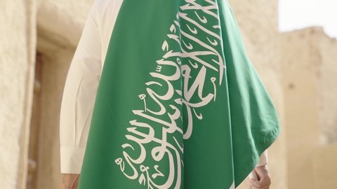 فن العمارة القديم وبناء البيوت الطينية، يوم العلم 11 مارس, إحياء ذكرى اليوم الوطني السعودي 23 سبتمبر, إيماءات الفخر و السرور بالانتماء للوطن, رجل عربي خليجي سعودي مبتسم يرتدي الشماغ و الثوب التقليدي يقف في بيت طيني ويحمل بيده علم المملكة، يوم بدينا 1727 م