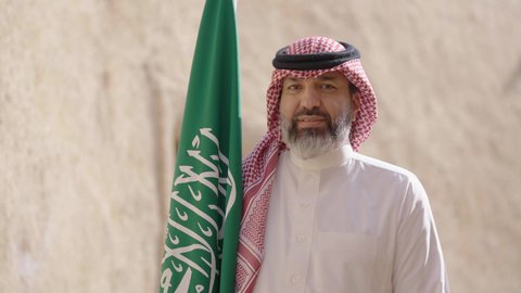 Ancient architecture and mud house construction, Flag Day March 11, Saudi National Day commemoration September 23, gestures of pride and joy in belonging to the homeland, a smiling Saudi Arabian Gulf man wearing a traditional shemagh and thobe standing in a mud house holding the Kingdom's flag, the day we began 1727 AD