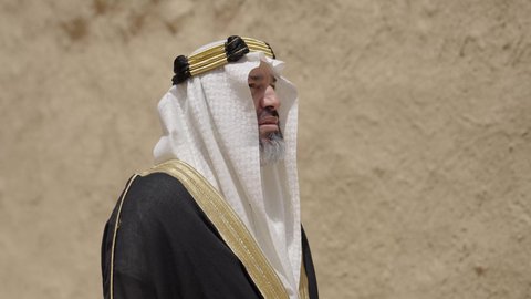 Commemorating the founding of the first Saudi state in 1727 AD, the day we began, February 22, a Saudi Gulf Arab man wearing a bisht, ghutra, and a wide golden agal stands in front of an old clay house, the concept of patriotism and belonging to the homeland, an old historical village, wearing traditional folk costume on a national holiday and occasion