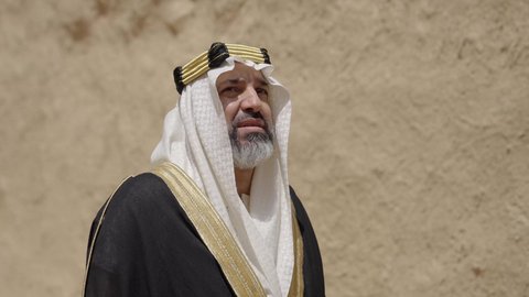 ارتداء الزي الشعبي  التقليدي في عيد ومناسبة وطنية، إحياء ذكرى تأسيس الدولة السعودية الأولى 1727 م, يوم بدينا 22 فبراير, رجل عربي خليجي سعودي يرتدي البشت والغترة والعقال الذهبي العريض يقف أمام منزل طيني قديم، مفهوم حب الوطن والانتماء اليه، قرية تاريخية قديمة