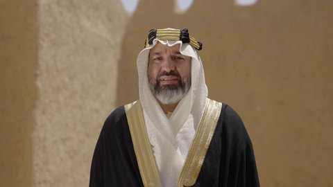 ارتداء الزي الشعبي  التقليدي في عيد ومناسبة وطنية، إحياء ذكرى تأسيس الدولة السعودية الأولى 1727 م, يوم بدينا 22 فبراير, رجل عربي خليجي سعودي يرتدي البشت والغترة والعقال الذهبي العريض يقف أمام منزل طيني قديم، مفهوم حب الوطن والانتماء اليه، قرية تاريخية قديمة