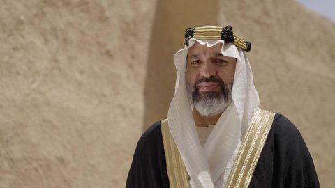ارتداء الزي الشعبي  التقليدي في عيد ومناسبة وطنية، إحياء ذكرى تأسيس الدولة السعودية الأولى 1727 م, يوم بدينا 22 فبراير, رجل عربي خليجي سعودي يرتدي البشت والغترة والعقال الذهبي العريض يقف أمام منزل طيني قديم، مفهوم حب الوطن والانتماء اليه، قرية تاريخية قديمة