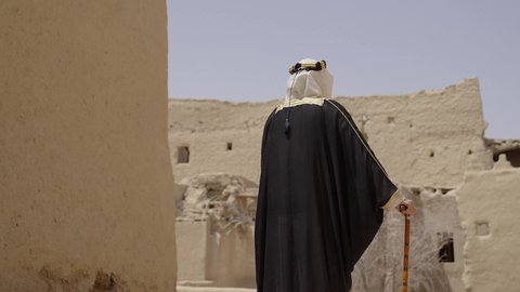 ارتداء الزي الشعبي  التقليدي في عيد ومناسبة وطنية، إحياء ذكرى تأسيس الدولة السعودية الأولى 1727 م, يوم بدينا 22 فبراير, رجل عربي خليجي سعودي يرتدي البشت والغترة والعقال الذهبي العريض يقف أمام منزل طيني قديم، مفهوم حب الوطن والانتماء اليه، قرية تاريخية قديمة