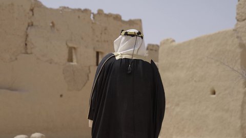 An ancient historical village, wearing traditional folk costume on a national holiday and occasion, commemorating the founding of the first Saudi state in 1727 AD, the day of our beginning, February 22, a Saudi Gulf Arab man wearing a bisht, ghutra and a wide golden agal standing in front of an old clay house, the concept of love of the homeland and belonging to it