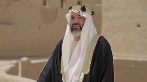 قرية تاريخية قديمة، ارتداء الزي الشعبي  التقليدي في عيد ومناسبة وطنية، إحياء ذكرى تأسيس الدولة السعودية الأولى 1727 م, يوم بدينا 22 فبراير, رجل عربي خليجي سعودي يرتدي البشت والغترة والعقال الذهبي العريض يقف أمام منزل طيني قديم، مفهوم حب الوطن والانتماء اليه