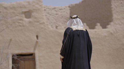 تعابير الفخر والاعتزاز،  قرية تاريخية قديمة، ارتداء الزي الشعبي  التقليدي في عيد ومناسبة وطنية، إحياء ذكرى تأسيس الدولة السعودية الأولى 1727 م, يوم بدينا 22 فبراير, رجل عربي خليجي سعودي يرتدي البشت والغترة والعقال الذهبي العريض يقف في منزل طيني قديم
